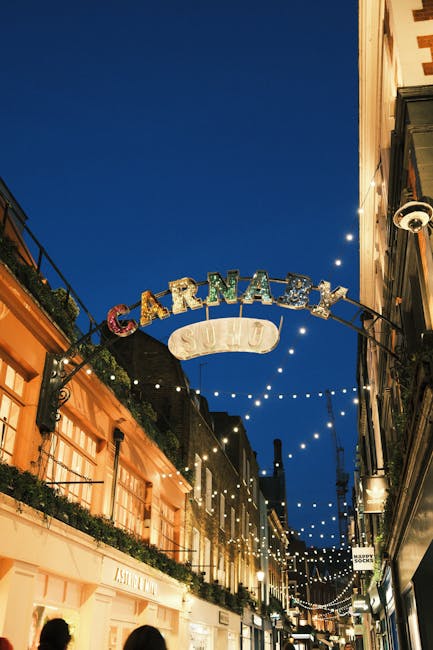 A nighttime street scene with a prominent illuminated sign reading 'CARNABY' hanging above a narrow pedestrian walkway, which is lined with shops and boutiques. The sky is dark blue, indicating evenin